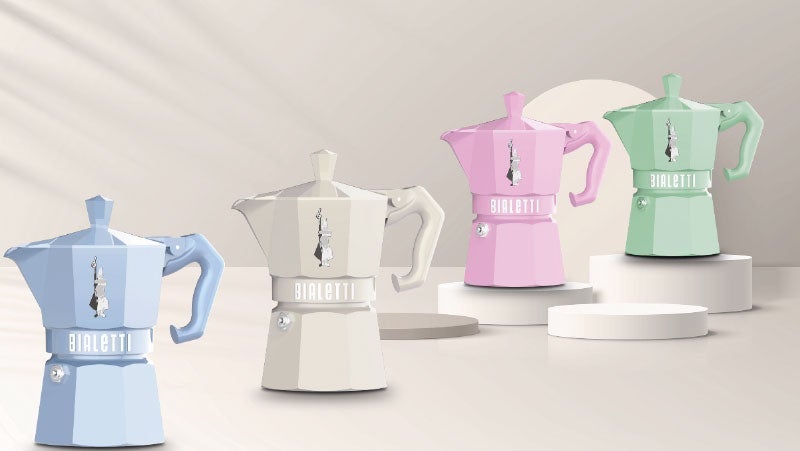 Assortment of Bialetti Moka pots in blue, beige, pink, and green, displayed on geometric platforms in a soft-lit setting