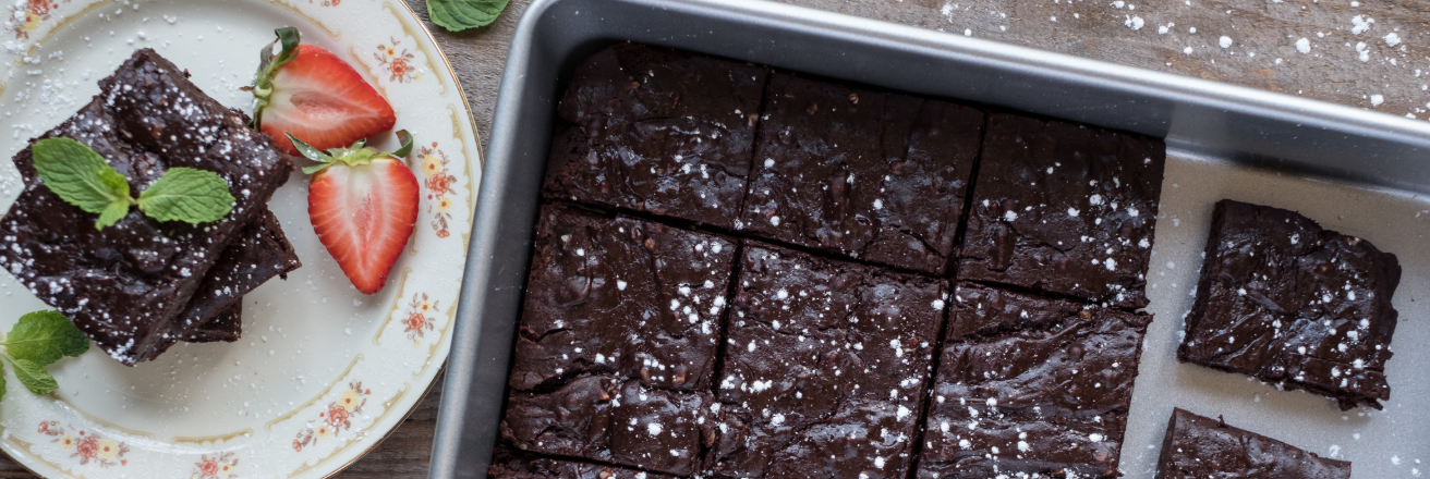 Chocolate cake with sugar in grey baking tray with plate and slice of cake with strawberries to the side