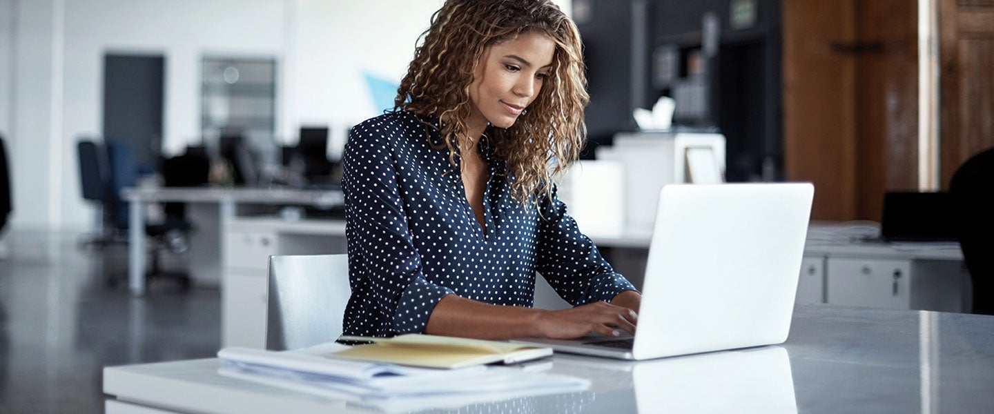 Professional woman with curly hair, wearing a blue polka dot blouse, working intently on a laptop at a sleek white desk in a bright, modern office environment.