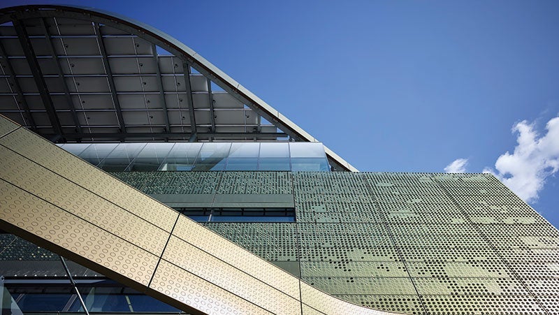 Low-angle view of a modern building featuring a curved roof and green perforated facade under a clear blue sky