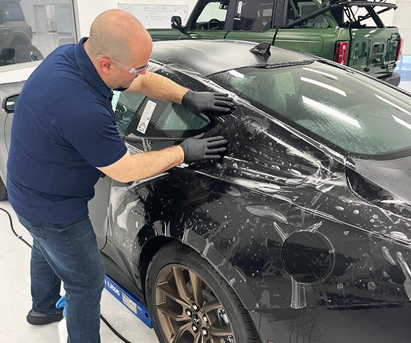 A man wearing goggles and gloves applying protective film to the body of a black car