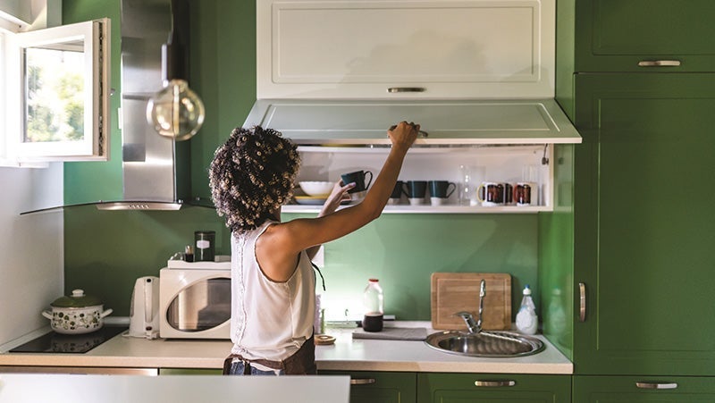 Woman wearing a white tank top organizing glassware in modern green kitchen cabinets