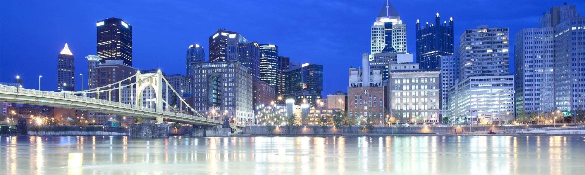 Pittsburgh skyline featuring the Roberto Clemente Bridge over the Allegheny River illuminated at dusk with reflections on the water