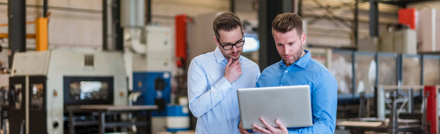 Two men in shirts in a manufacturing environment looking at a silver laptop screen