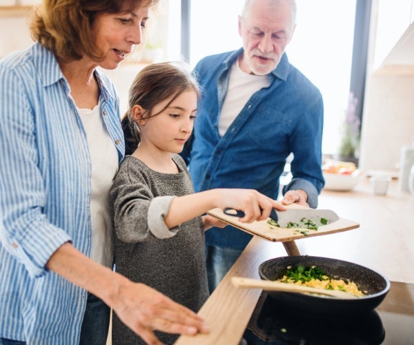 Family cooking together, daughter scraping cut greens into a black non-stick frying pan on a cooker hob