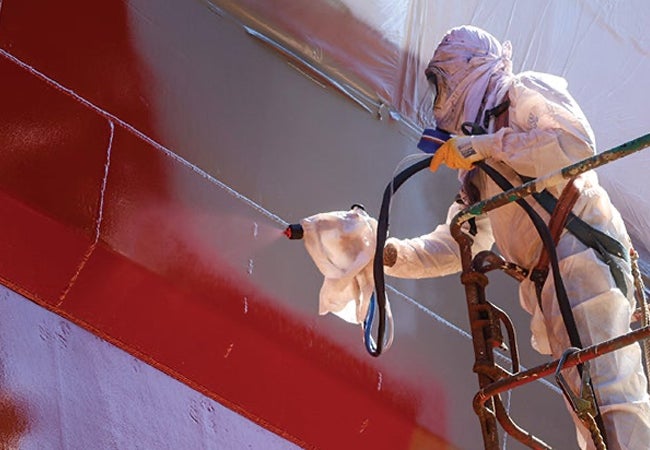 Worker wearing PPE spraying ship with red coating