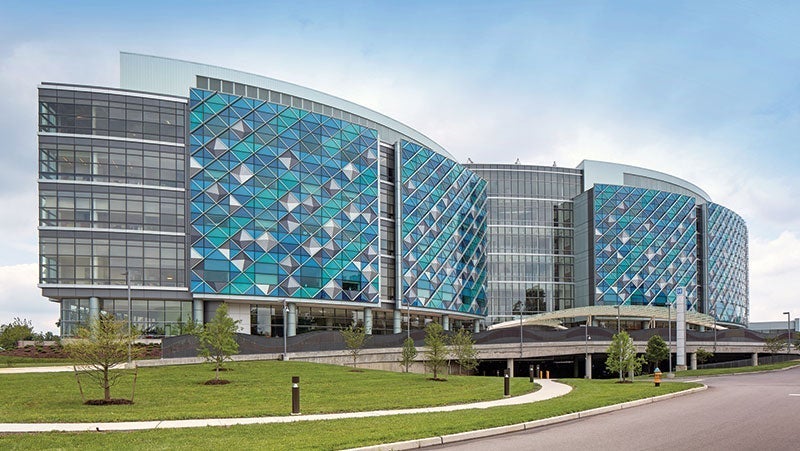 Contemporary building with blue patterned glass facade and curved design, featuring an overpass and landscaped surroundings