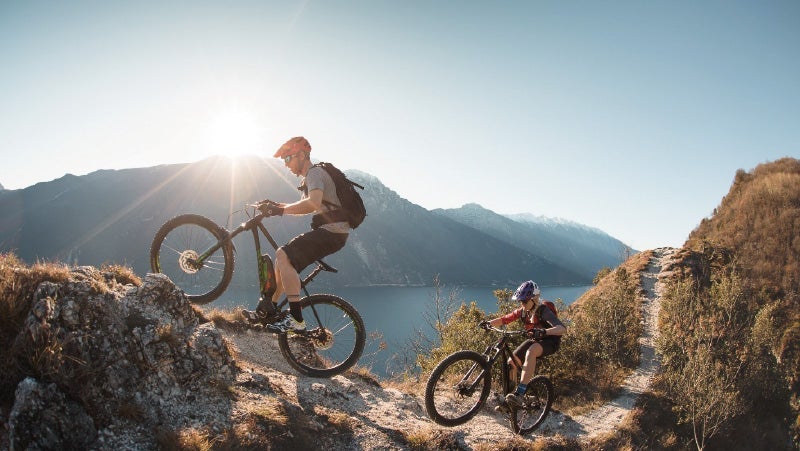 Two mountain bikers navigate a rugged trail above a serene lake in bright sunlight