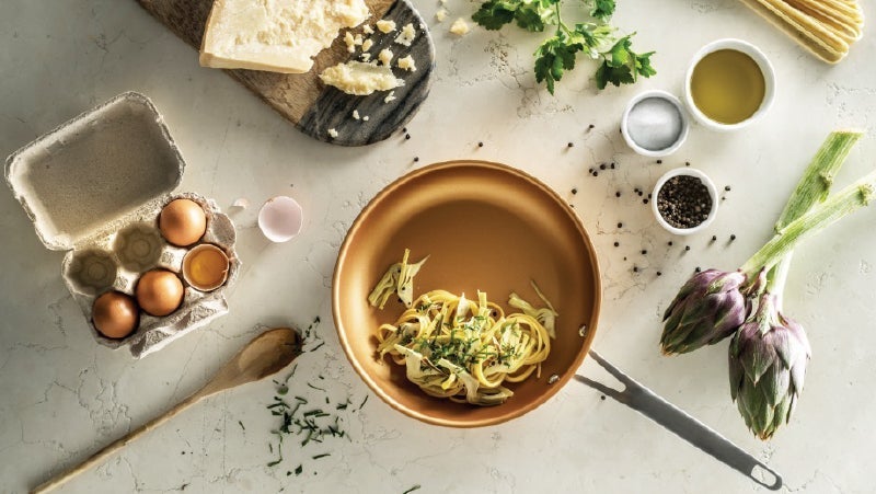 Top view of gourmet cooking scene with fresh ingredients: eggs in carton, olive oil, herbs, parmesan cheese, pepper, and artichokes on a marble countertop