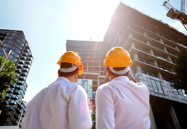 Two architects wearing yellow hard hats ling up at a tall building