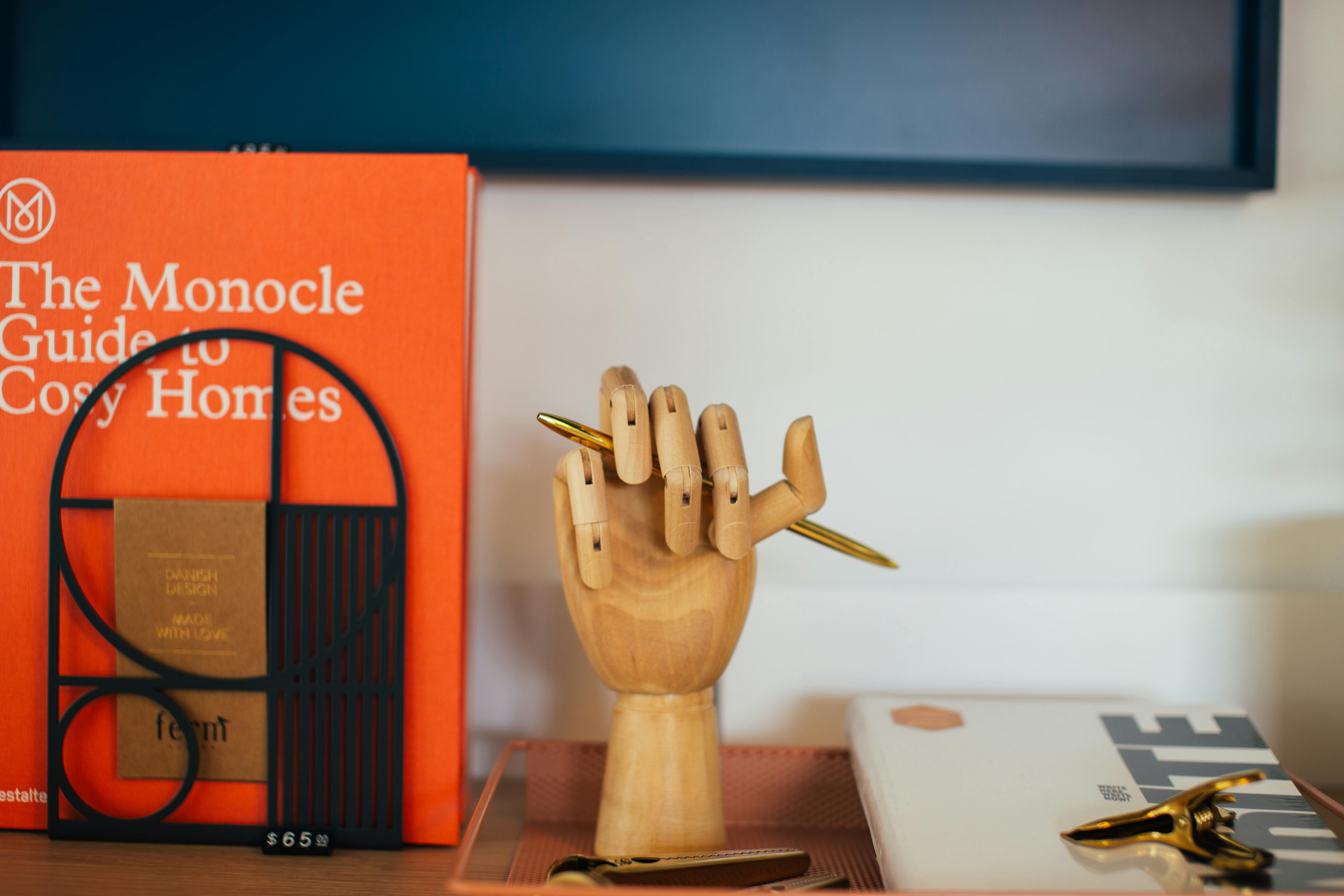 Wooden hand holding a pencil in front of an orange book - The Monocle Guide to Cosy Homes