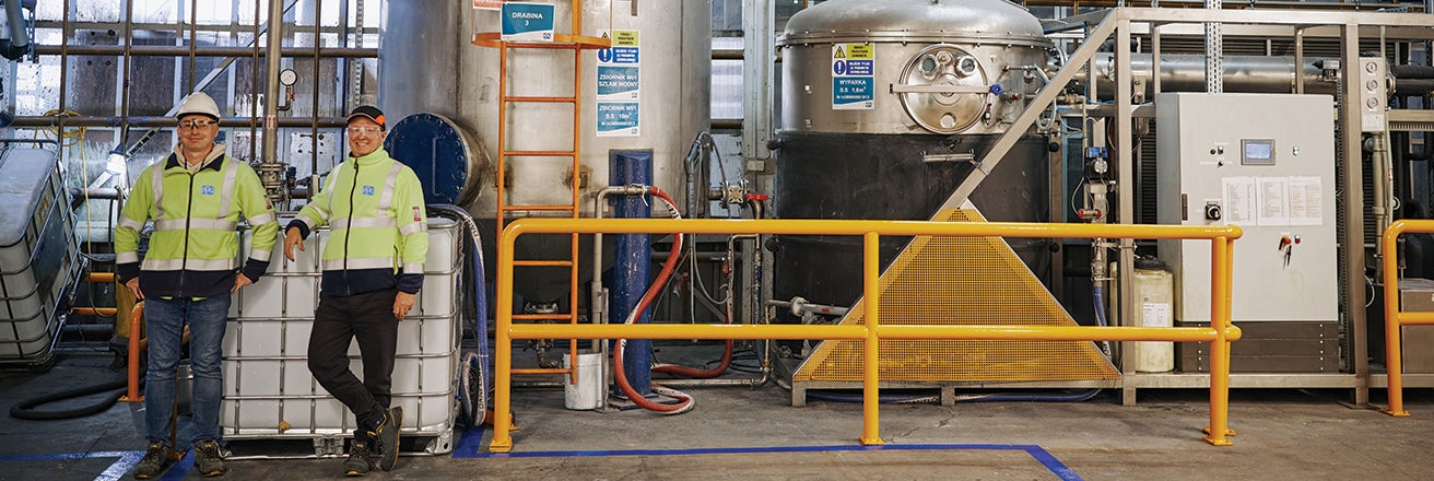 Two male PPG workers in a factory alongside large containers and pipework