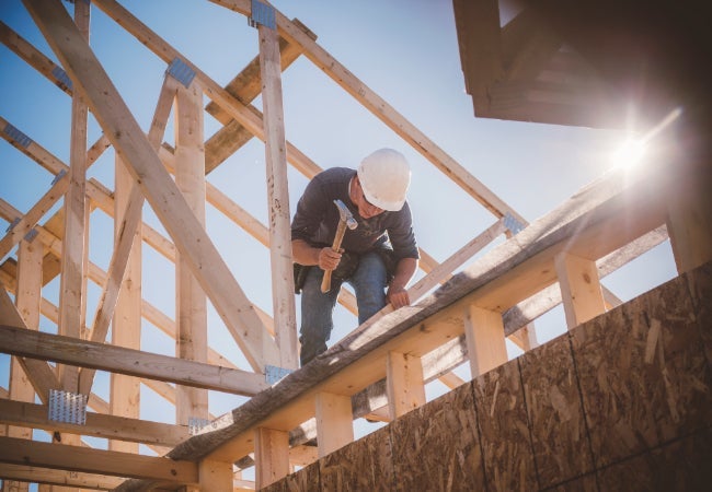 Construction worker in hard hat using a hammer on a wooden beam at a sunny construction site