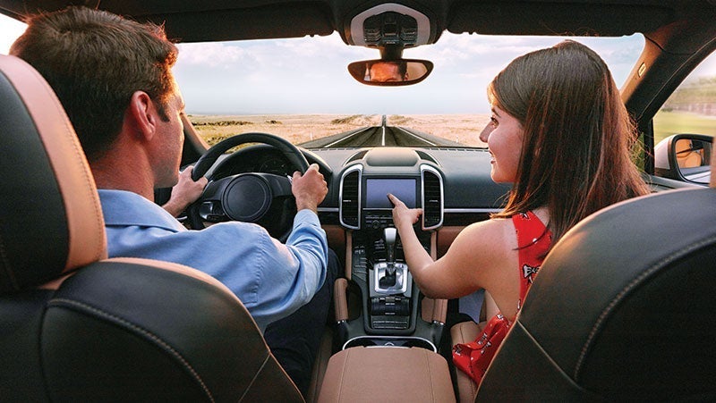 Rear view of a couple driving in a luxury car on a scenic road at sunset, man holding the steering wheel while woman gestures to the dashboard