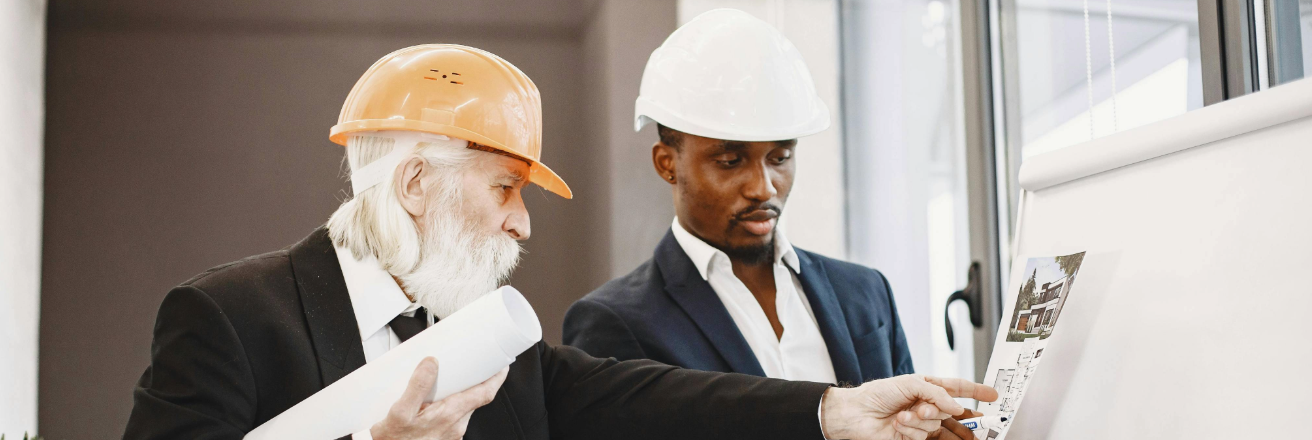 Two architects, one with a grey beard wearing an orange hard hat, the second wearing a white hard hat, looking at building plans