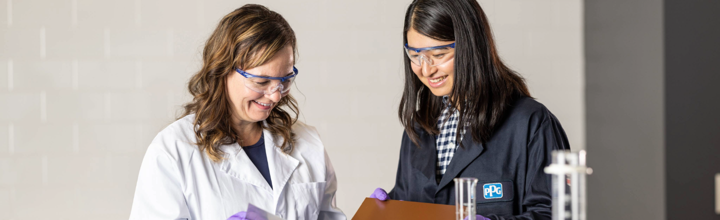 Two women wearing safety glasses and lab coats looking at test data