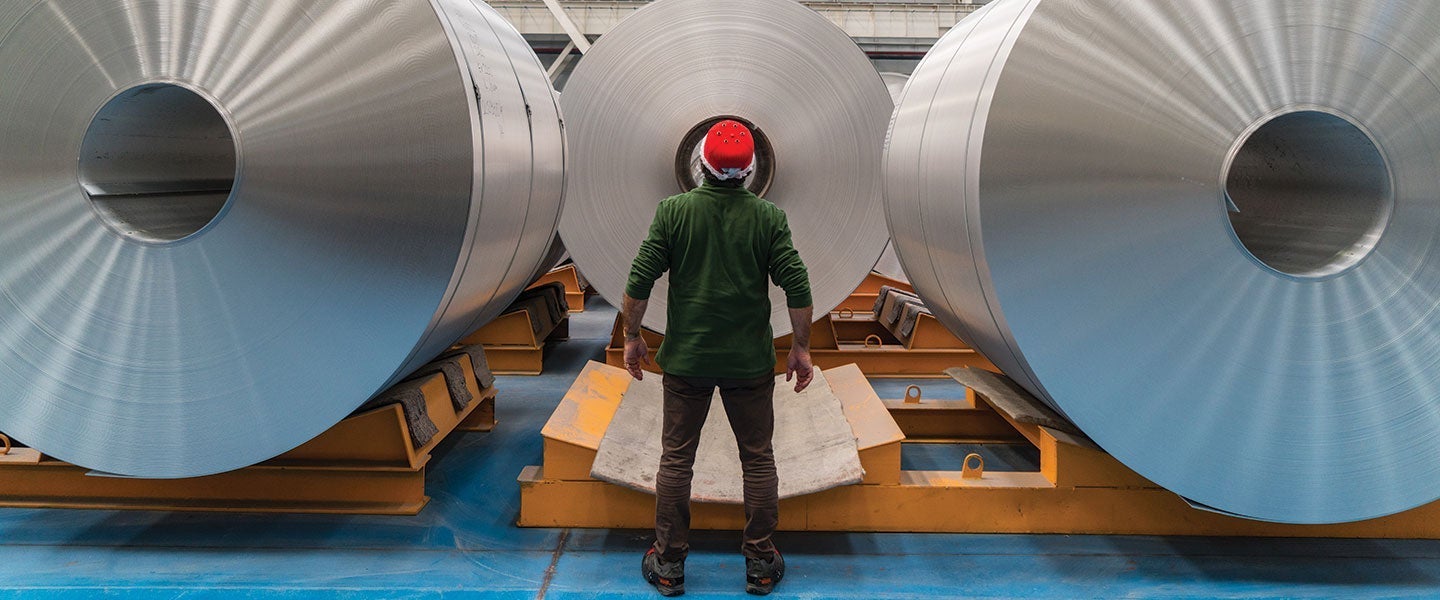 Back view of a worker wearing a red helmet and green jacket standing in front of large aluminum coils in an industrial setting