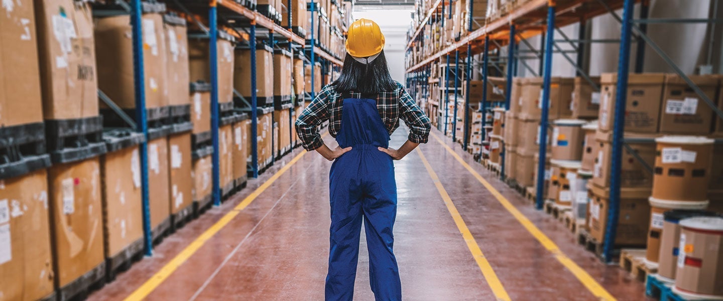 Confident female warehouse worker in a hard hat standing with hands on hips in a warehouse aisle filled with high shelves of cardboard boxes