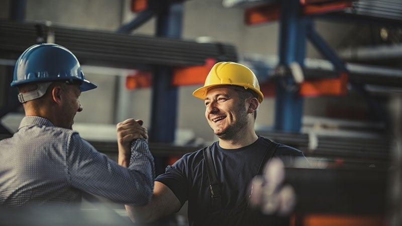 Two industrial workers inspect machinery in a factory, one wearing a blue hard hat and the other in a yellow hard hat, focused on their teamwork.