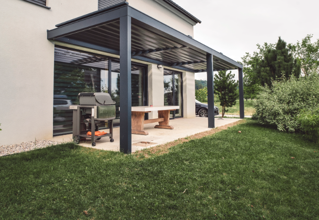 Contemporary white house with a stylish metal pergola covering a wooden dining table and barbecue grill, surrounded by green lawn and bushes