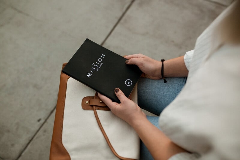 Photo by David Iskander on Unsplash, a female sitting down with a handbag on her lap holding a black book with the words 'THE MISSION JOURNAL' on it
