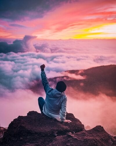 Photo by Ian Stauffer on Unsplash, a man sitting on some large rocks with one arm in the air surrounded by clouds