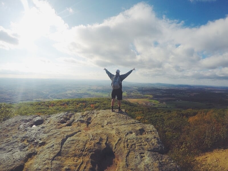 Photo by Jake Ingle on Unsplash, a man standing on top on a hill with his arms in the air, wearing a rucksack, baseball hat and shorts. 