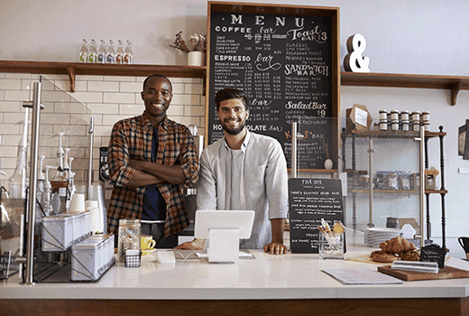 Two small business owners stand behind the counter and in front of a large menu sign at their coffee shop