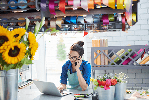 A business owners sits in front of a laptop and speaks on her phone. She has sunflowers on her desk and textile and ribbons in her workplace