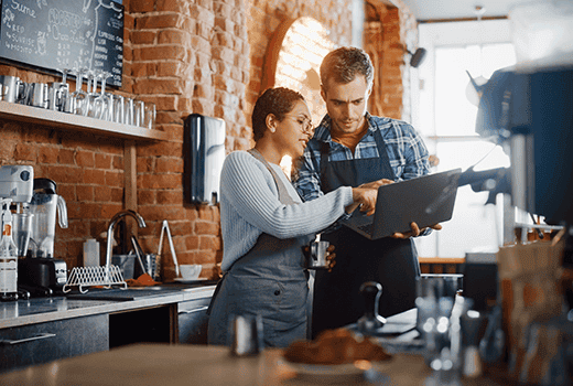 An employer shows an employee something on a laptop behind the counter of her coffee shop