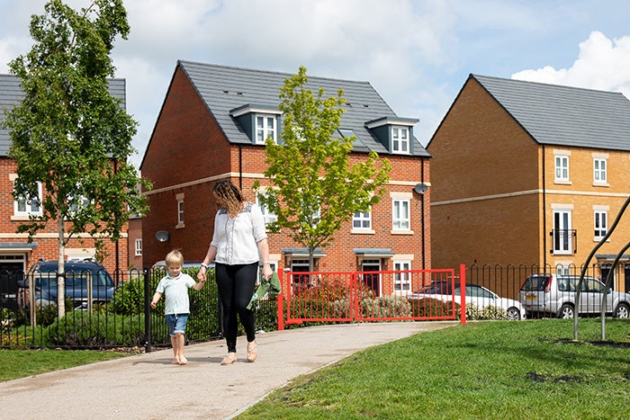 A mother and child walking through a playground, with some red brick homes behind them