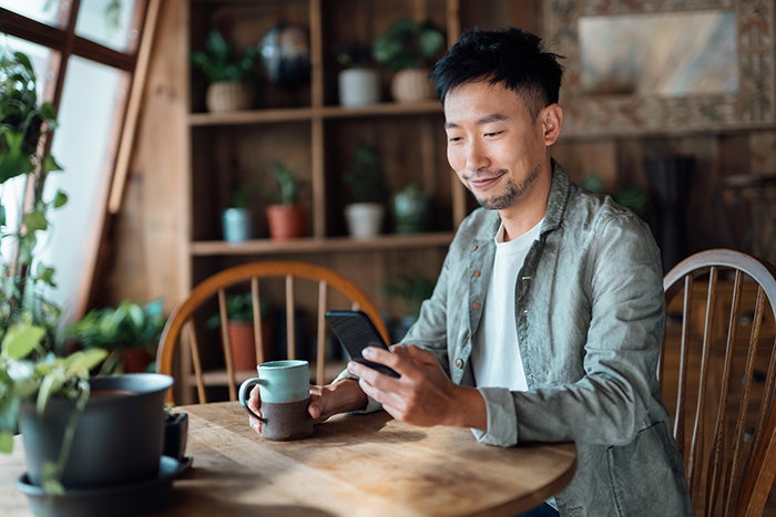 A man sitting at his kitchen table with a cup of coffee in one hand and his mobile phone in the other