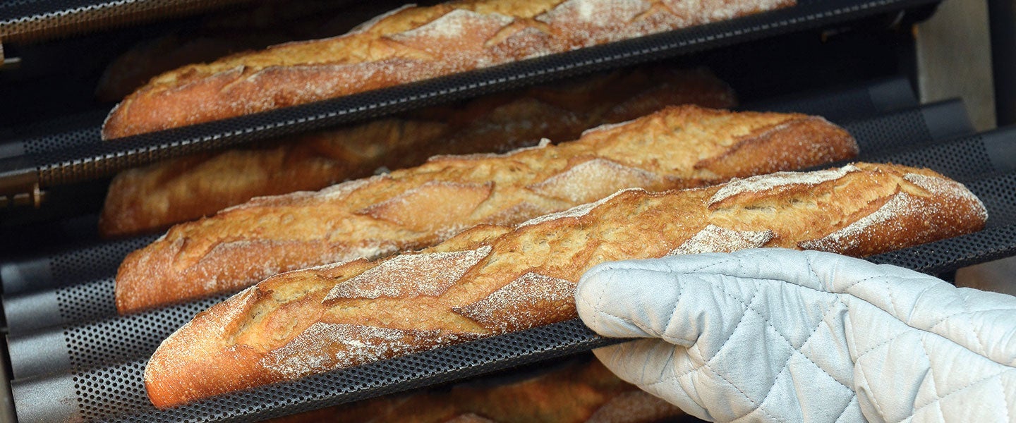 Close-up view of a hand wearing an oven mitt removing freshly baked baguettes from a professional oven