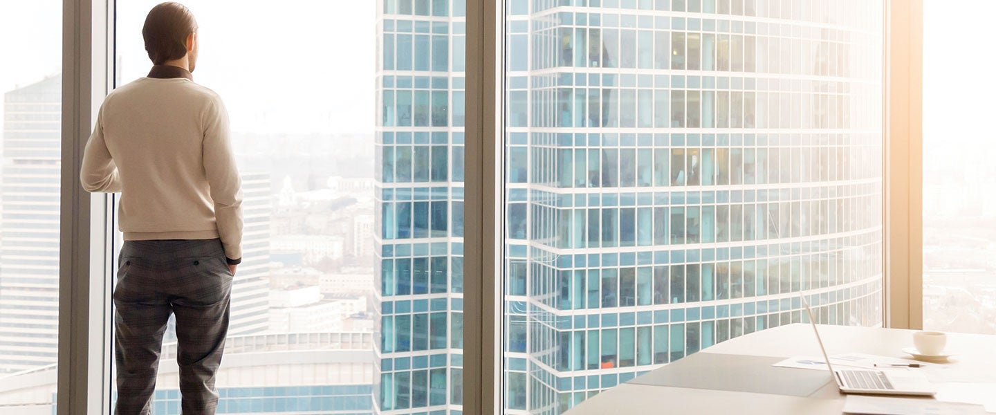 Man in business casual attire standing by a large window in a modern office, looking out at a sprawling urban cityscape with skyscrapers