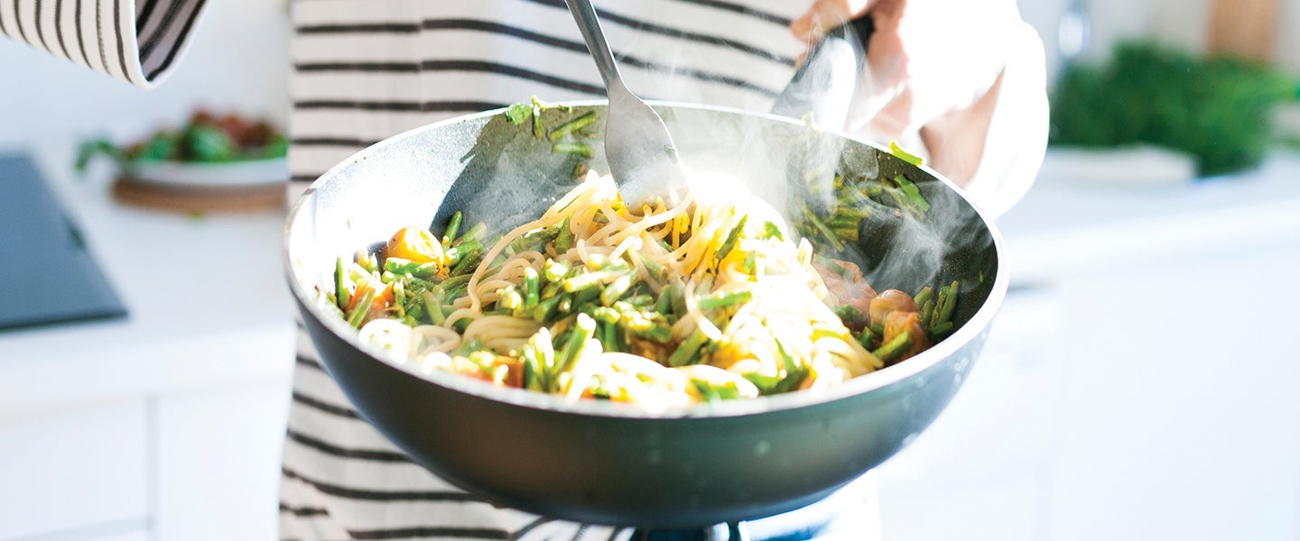 Person in striped shirt cooking vegetable stir-fry with noodles in a modern kitchen, steam rising from the skillet