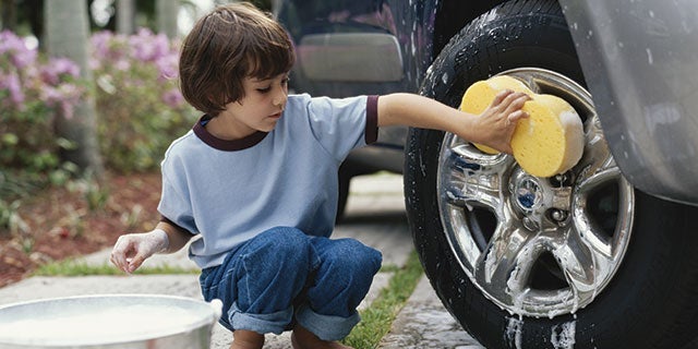 Young child in a blue shirt and jeans kneeling while cleaning a car wheel with a yellow sponge, next to a bucket of soapy water