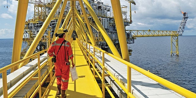 Worker in red safety gear walking along a yellow metal walkway on an offshore oil rig, with ocean and rig structures in the background