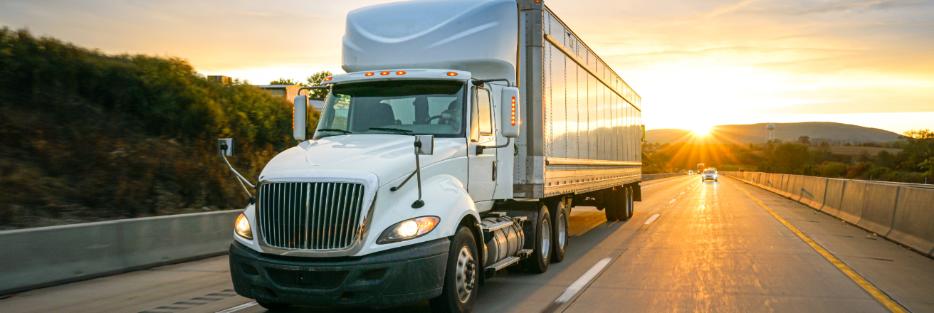 White truck with a trailer carrying a large container driving down the highway against a sunset sky