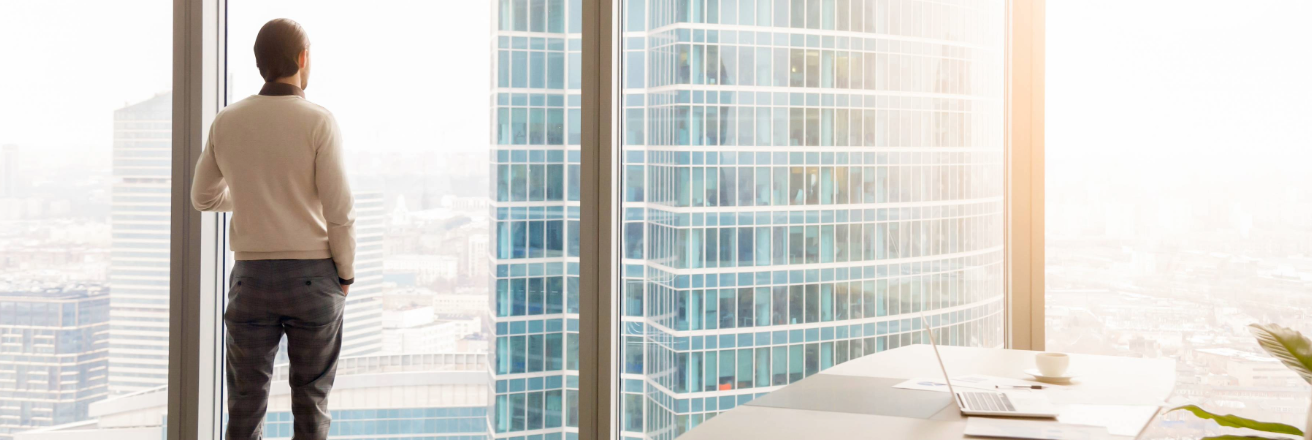 Man in business casual attire standing by a large window in a modern office, looking out at a sprawling urban cityscape with skyscrapers