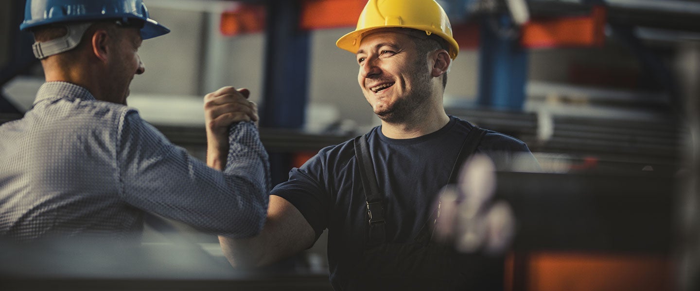 Two industrial workers in a manufacturing plant, one wearing a blue hard hat and the other in a yellow hard hat, collaborating over machinery