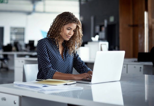 Woman in a polka dot blouse working on a laptop at a clutter-free desk in a bright, spacious office.