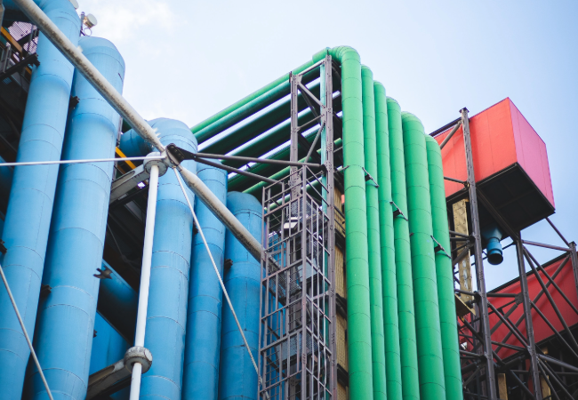 Large green and blue pipes in a refinery against a red storage area