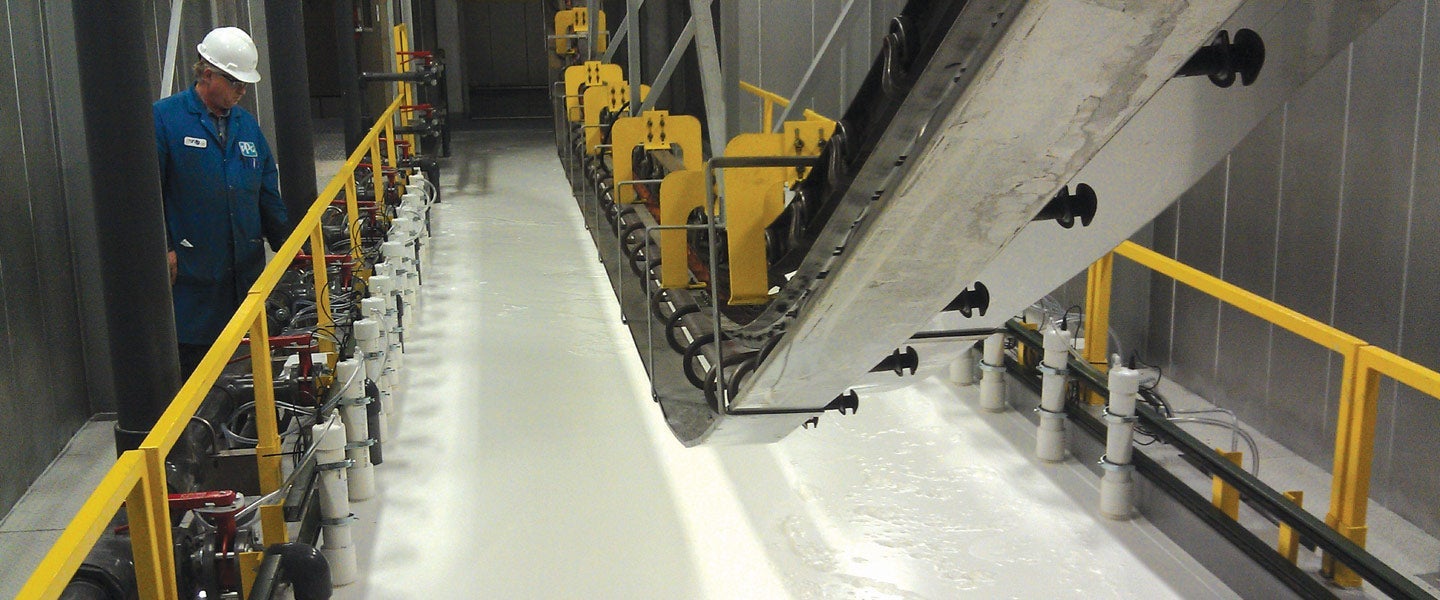 Worker in a blue uniform inspects a large conveyor belt mechanism in an industrial setting with yellow safety barriers and intricate piping on the floor.