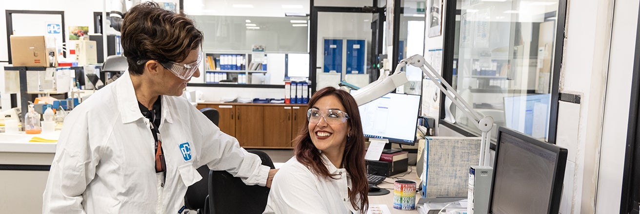 Two female PPG workers wearing PPE discussing next to computer