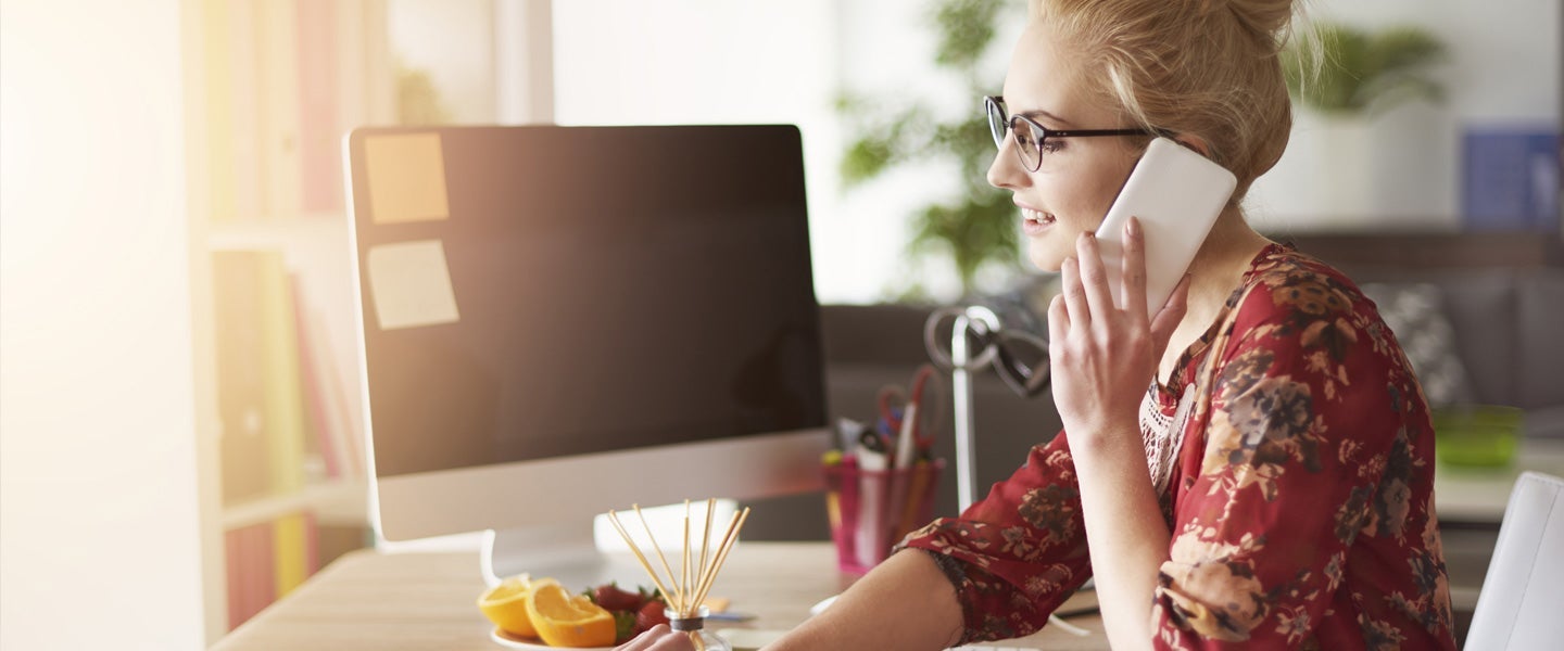 Woman in a floral blouse working at a modern workstation with a large monitor, speaking on the phone in a bright office space