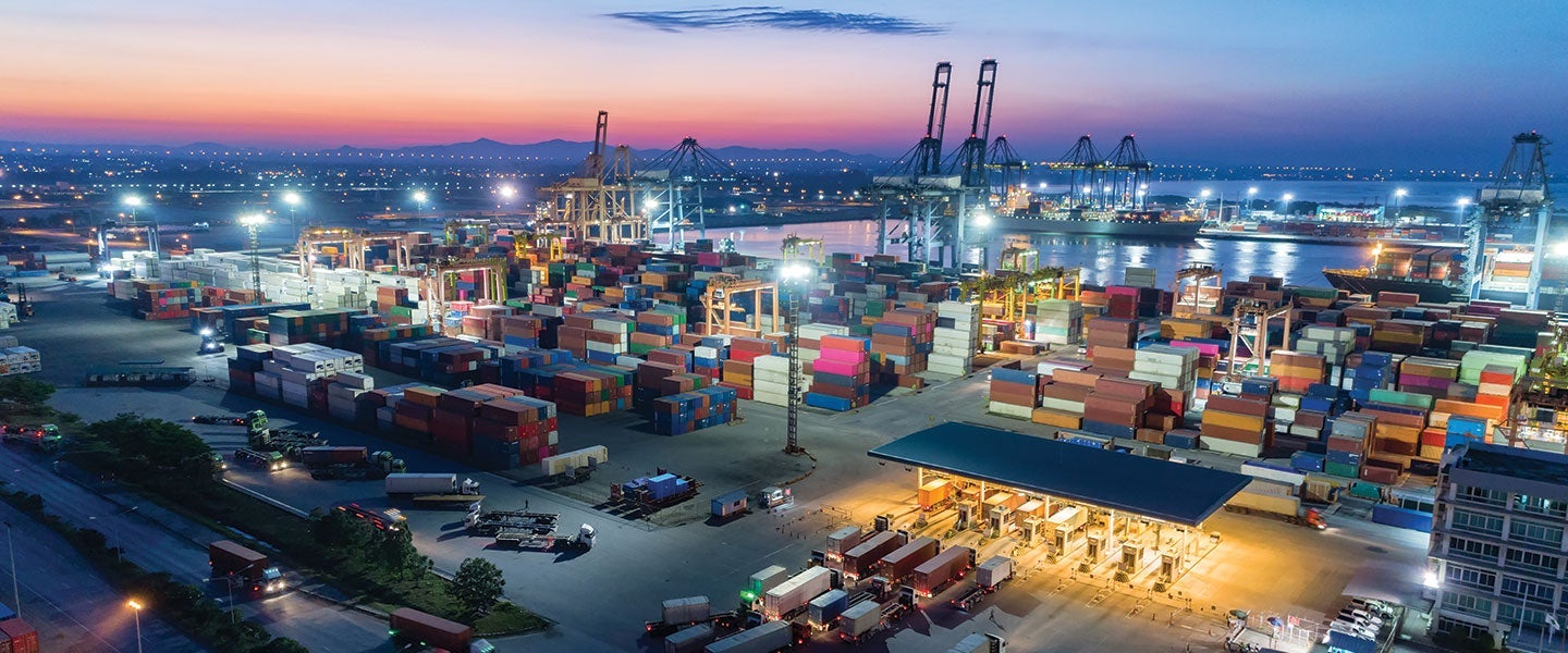 Aerial view of a bustling container port at twilight with colorful shipping containers, cargo trucks and cranes, with a backdrop of a fading sunset and distant mountains