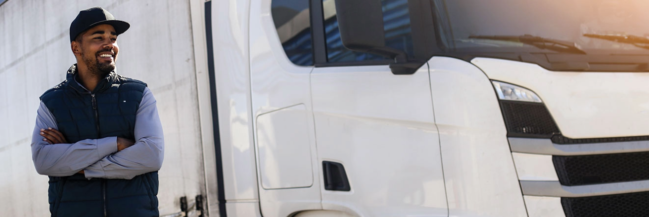 Truck driver with arms crossed standing confidently next to a white semi-truck in a sunny parking area