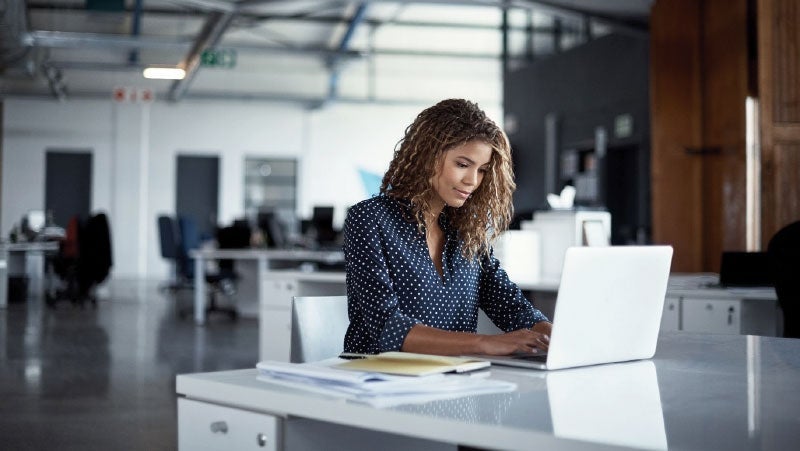 Professional woman focused on her laptop in a modern office setting with open desks and minimal design