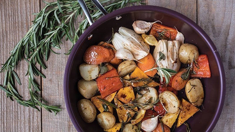 Colorful roasted vegetables in a purple bowl garnished with fresh rosemary, featuring carrots, potatoes, pumpkin, onions, and garlic cloves