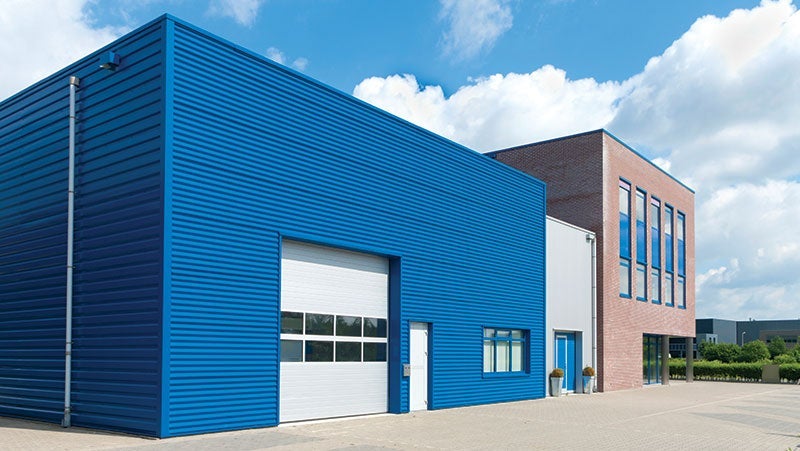 Exterior view of a modern industrial building featuring blue corrugated metal walls, large white garage doors, and an attached brick office section under a clear sky